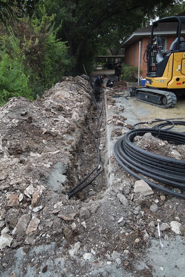 Open trench with geothermal loop piping running toward the house, mini excavator behind it